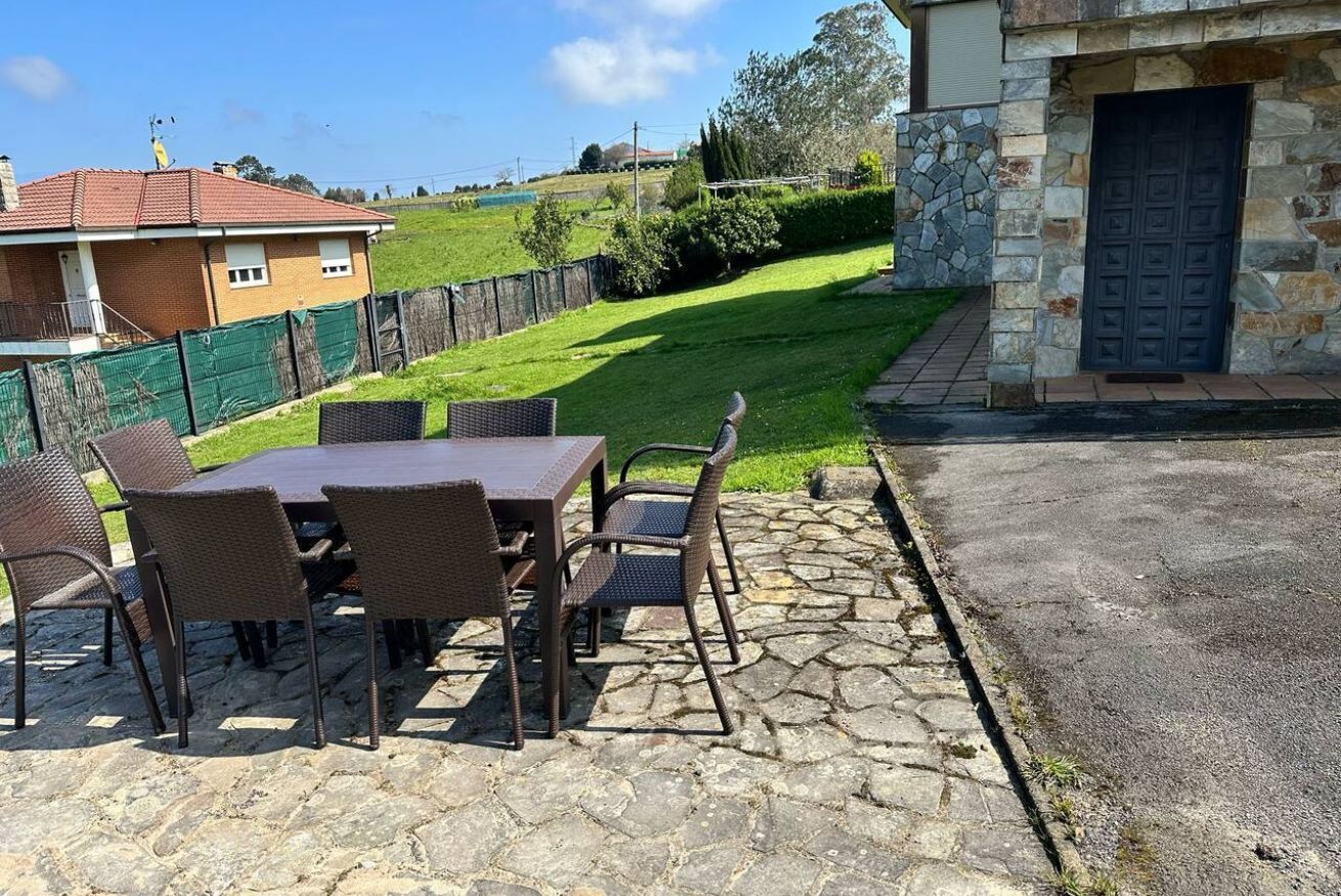 Eine Terrasse im Freien mit einem Steintisch und Korbstühlen, einer Steinsäule und Blick auf eine grüne Rasenfläche und ein Haus.