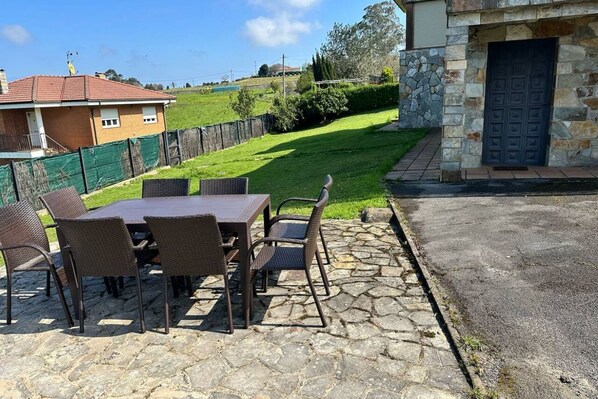 Un patio al aire libre con una mesa de piedra y sillas de mimbre, un muro de piedra y vistas a un césped verde y una casa.