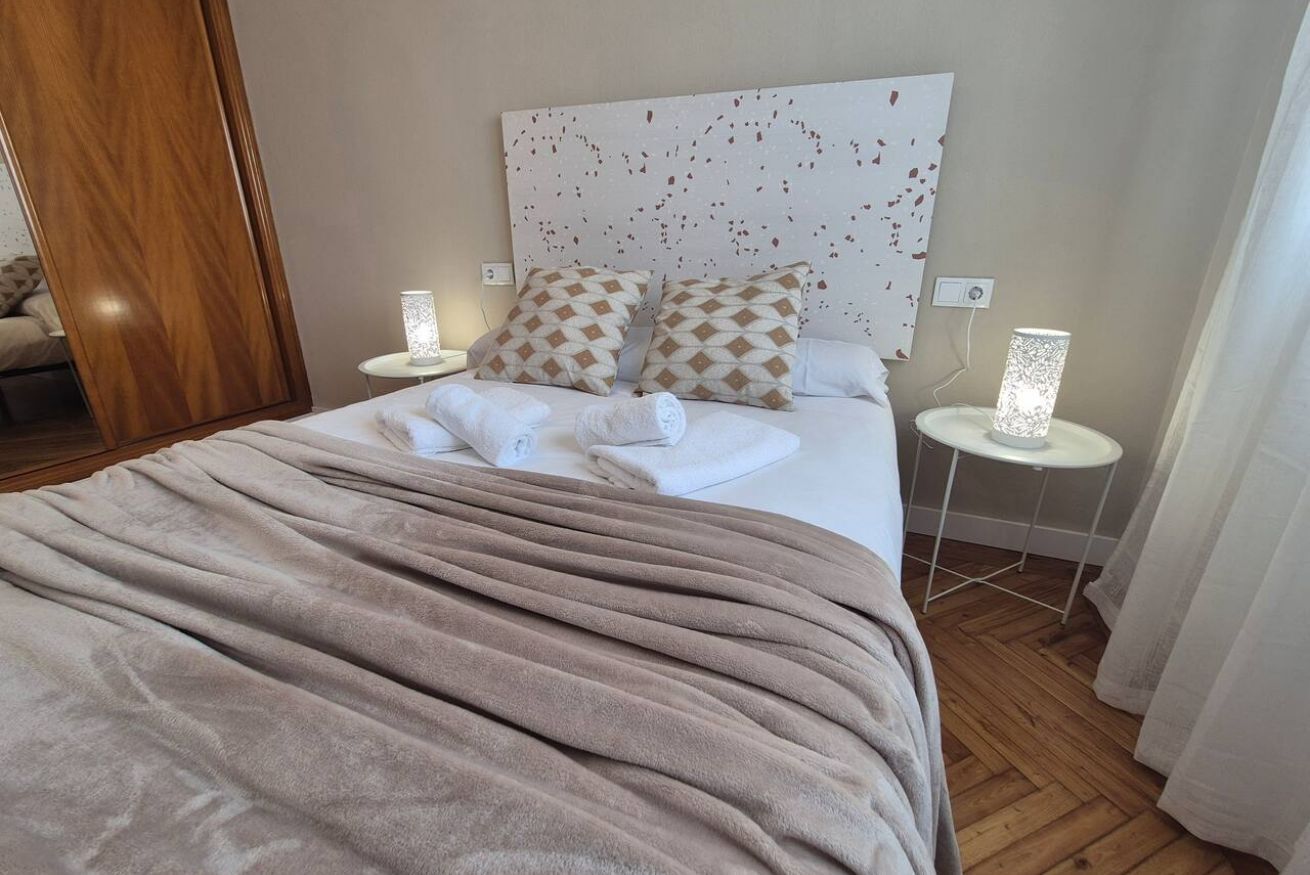 A neatly made bed with a beige blanket, two patterned pillows, and two bedside lamps on white nightstands.