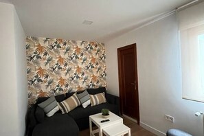 A living room with a sofa, a small table with a plant, and a wall with a tropical leaf pattern. - 803A Bonito apartamento en Luarca (Valdés)