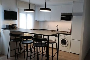 A modern kitchen with a central island, bar stools, a washing machine, and a flat-screen TV mounted on the wall.