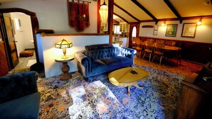 A cozy room with a blue sofa, a wooden coffee table, and a patterned carpet. There is a dining area with chairs and a table visible in the background.