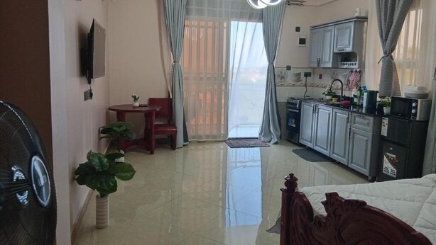 A compact hotel room with a bed, a small dining table, a kitchenette, and a large window with curtains. - Stunning Studio Apartment on Entebbe that is fully furnished and accessible (Namulanda)
