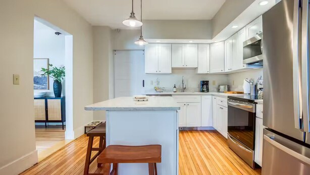 A modern kitchen with white cabinets, a central island, stainless steel appliances, and wooden flooring. - Home in Heart of Downtown North Conway (Conway)