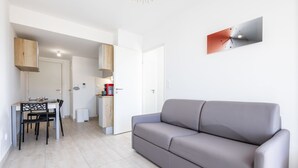 A modern living room with a grey sofa, a dining table with chairs, a kitchen area, and a wall-mounted clock.