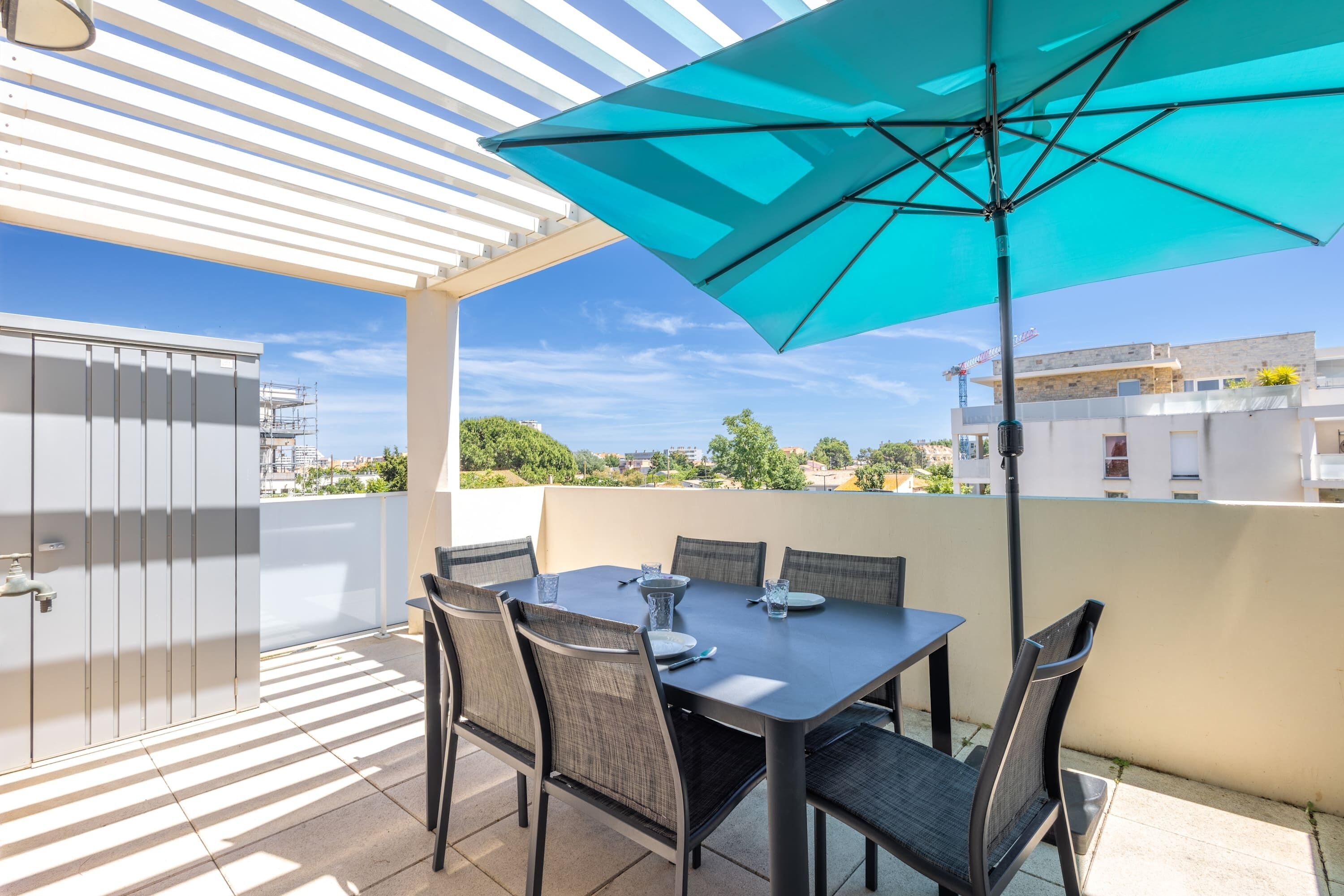 Une terrasse extérieure couverte, avec une table et des chaises, un parasol bleu et une vue sur des bâtiments et de la verdure.