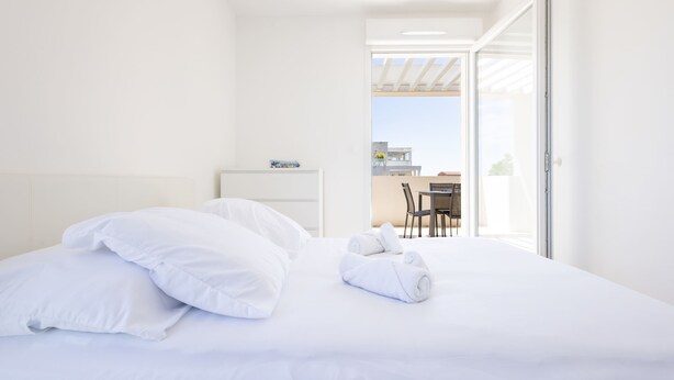 A hotel room with a bed, pillows, a rolled towel, a small table with a chair, and a view through an open door.