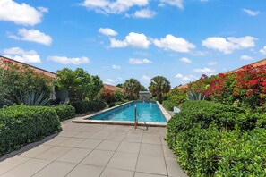 A rectangular swimming pool surrounded by lush greenery and red flowering plants, with a clear blue sky above.