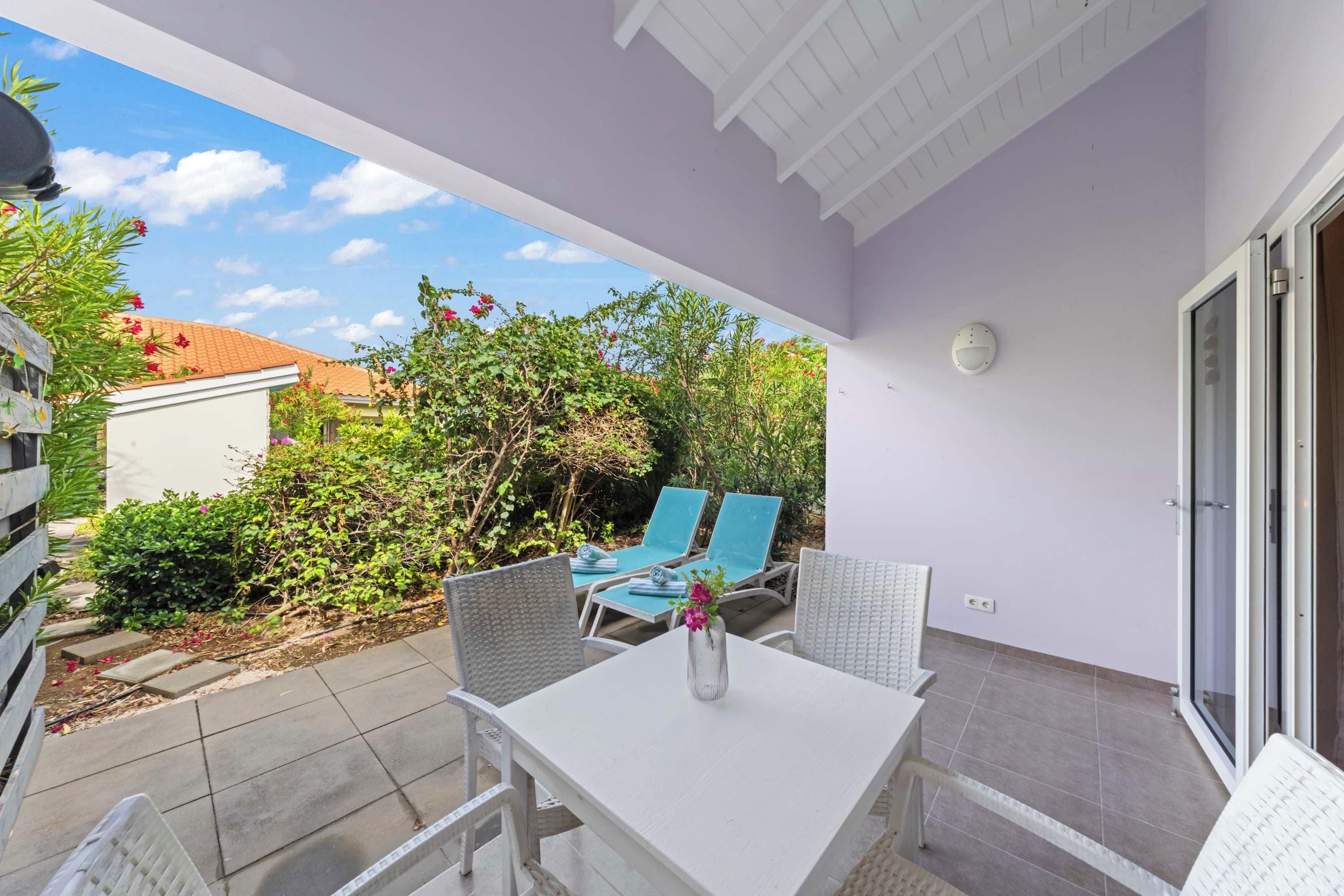 A patio with a table and chairs, a view of the garden, and a white building in the background.