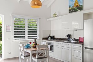 A modern kitchen with white cabinetry, a dining table with chairs, a hanging pendant light, and a window with blinds.