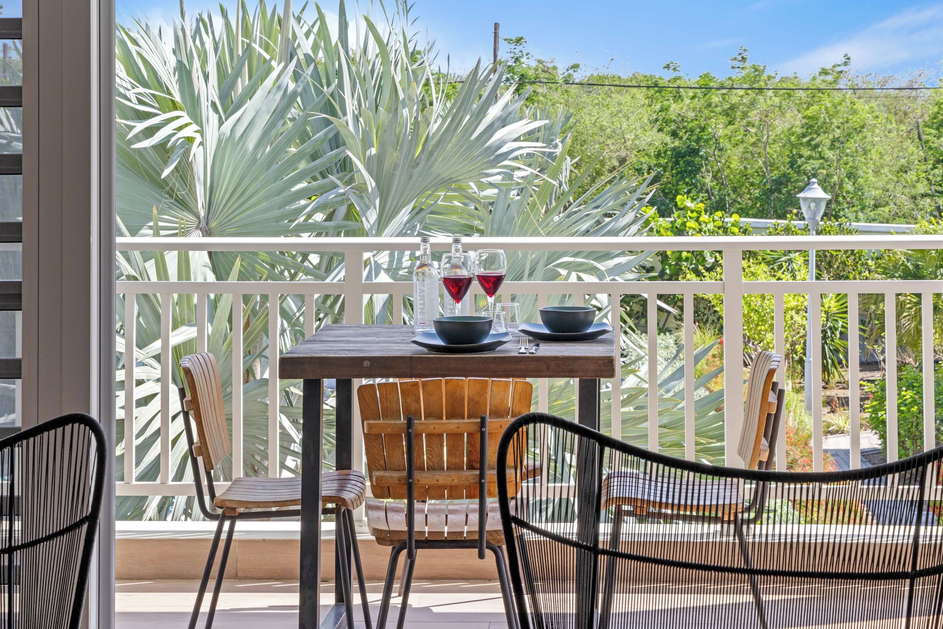 A balcony with a table set for two, two chairs, and a view of a garden with large white plants.
