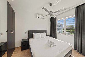 A modern hotel room with a large bed, a ceiling fan, an air conditioning unit, and a window with a view of buildings and greenery.