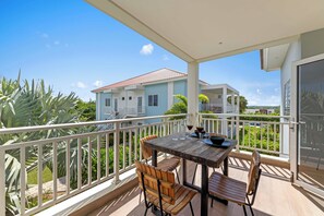 A balcony with a wooden table and chairs, a glass railing, and a view of a light blue house with a red roof.