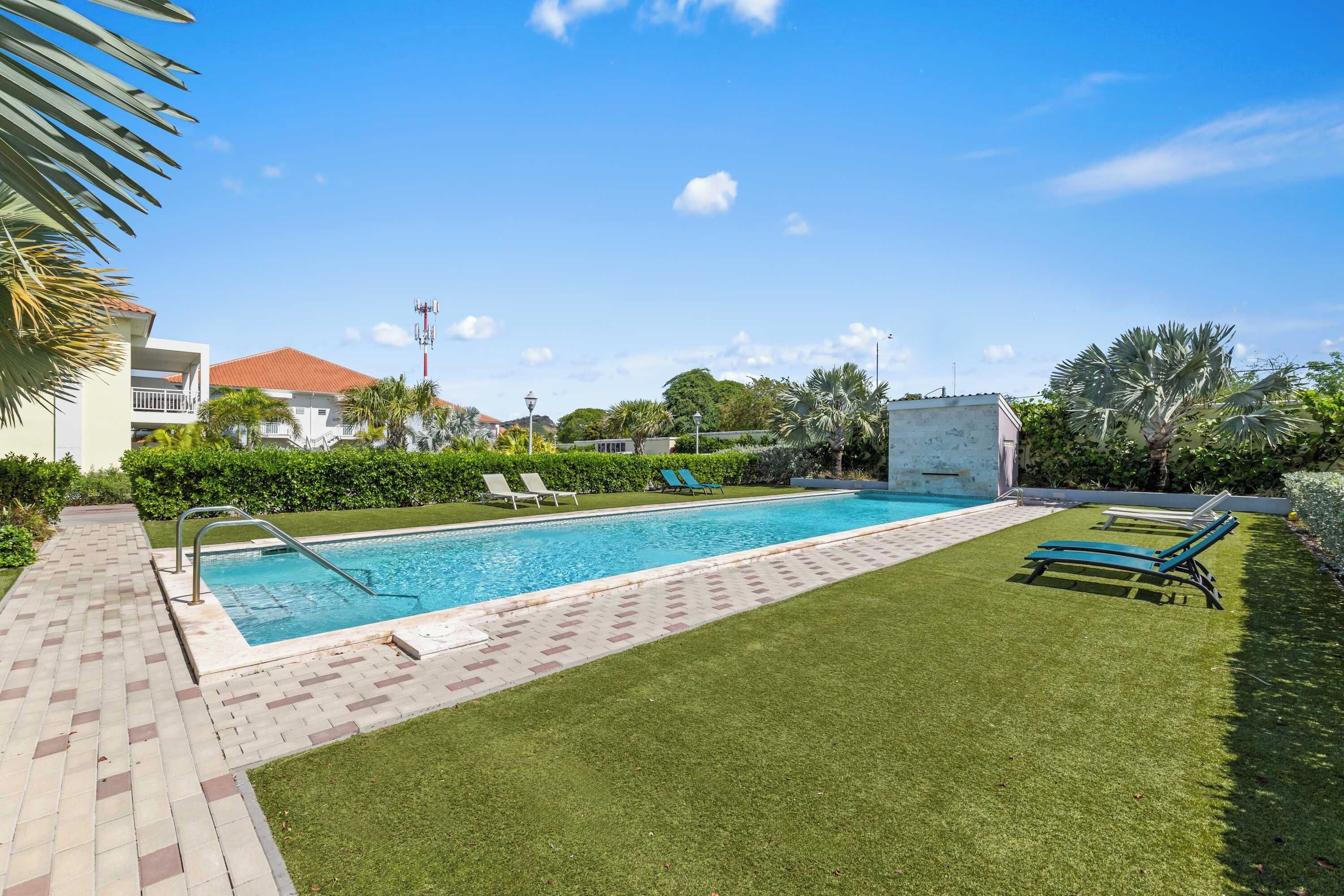 Una piscina con solárium empedrado, rodeada de césped verde y palmeras.