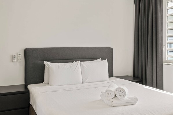 Una cama bien hecha con sábanas y almohadas blancas, un cabecero oscuro y un juego de toallas dobladas sobre la cama.