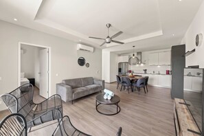 A modern living room with a grey sofa, black wireframe chairs, and a round coffee table. A ceiling fan and an air conditioner are visible. The kitchen area is in the background with white cabinets and a dining table.