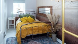 Un dormitorio con estructura de cama metálica, una mesita de noche con lámpara, un espejo en la pared y una planta decorativa.
