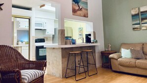 A modern kitchen with white cabinets, a built-in microwave, and a coffee machine. There is a wicker chair and a sofa in the living area.