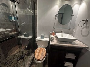 Un baño moderno con ducha de vidrio, asiento de inodoro de madera, lavamanos con encimera de mármol y espejo redondo.