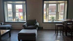 Una habitación con sofá, reposapiés, mesa de comedor, sillas, dos ventanas con vistas a edificios y una planta en maceta.