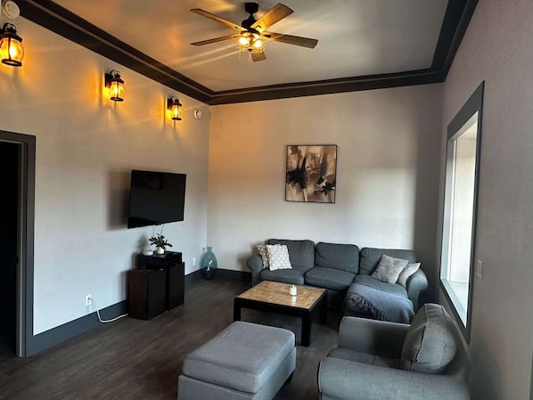 Un salon moderne avec un canapé d’angle, une table basse, un téléviseur fixé au mur et un ventilateur de plafond.