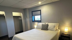 Una cama bien hecha con colcha, una lámpara de noche, una ventana con persianas y un perchero.