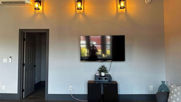 Un salón con una televisión de pantalla plana montada en la pared, suelo de madera oscura, un sofá gris y un mueble de entretenimiento negro pequeño.