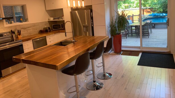 Une cuisine moderne dotée d’un îlot central en bois, d’appareils électroménagers en acier inoxydable et d’une grande fenêtre donnant sur un jardin avec piscine.