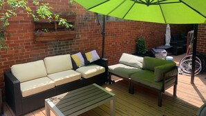 Une terrasse avec un mur en briques, un parasol vert, un canapé en osier noir, une chaise verte et une petite table.