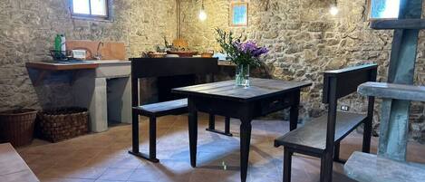 A rustic stone-walled room with a wooden table and benches, a stone fireplace, and a window.