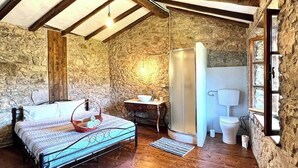 A rustic bedroom with a stone wall, wooden beams, a bed with a basket, a small table, and a toilet. - Tuscany Escape (Toscana)