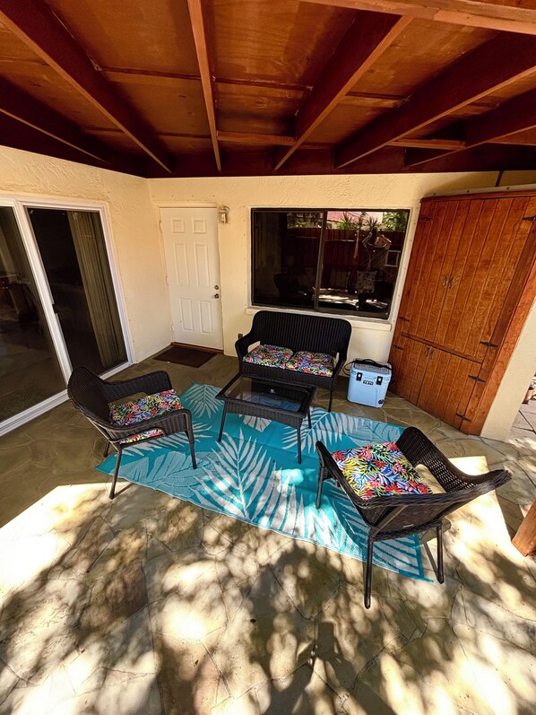 Un patio techado con mobiliario de mimbre, alfombra azul estampada y techo de madera.
