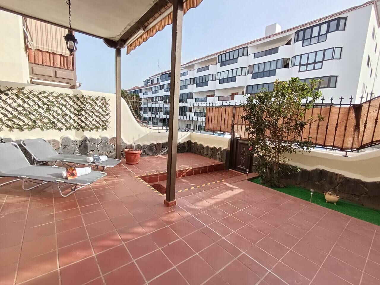 A covered outdoor area with tiled flooring, lounge chairs, and a small garden.