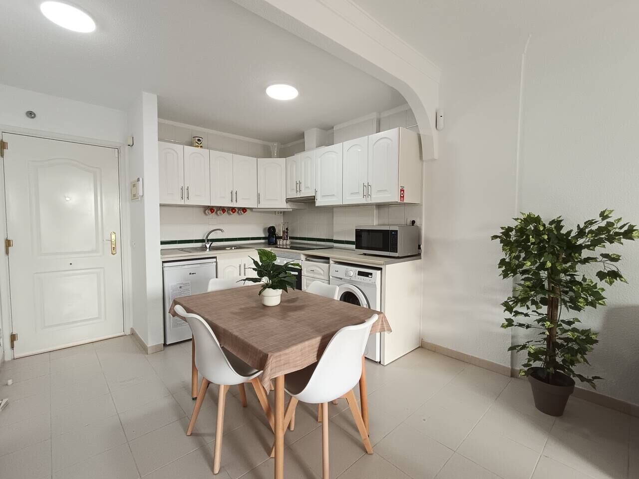 A compact kitchen with white cabinets, a microwave, a washing machine, and a dining table with chairs.