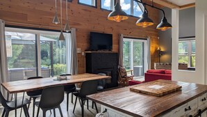 A modern kitchen with a wooden island, dining table, and chairs. There is a fireplace, large windows, and a TV mounted on the wall. - Cozy River Lodge Chalet - AC - Hot Tub - Waterfront - Pet Friendly (Saint-Côme)