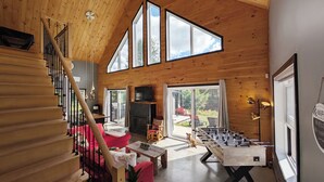 A spacious living room with a high ceiling, wooden walls, a fireplace, a foosball table, and a large window offering a view of the outdoors. - Cozy River Lodge Chalet - AC - Hot Tub - Waterfront - Pet Friendly (Saint-Côme)