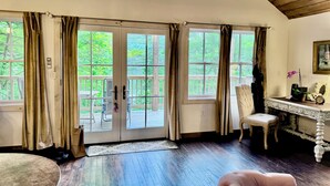 A room with a wooden ceiling, a round skylight, a large window with curtains, a white chair, a small table with a clock, and a framed picture on the wall. - Lakeview Cottage (Lake Hill)
