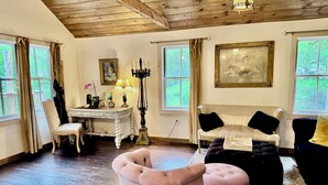 Ein Zimmer mit Holzboden, einem weißen, verzierten Tisch, einer weißen Couch mit schwarzen Kissen, einem pinkfarbenen Sessel und einem großen Gemälde an der Wand.
