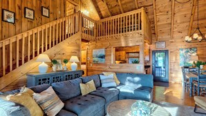 A wooden cabin interior with a staircase, a sofa, a dining area, and a sign indicating cabin rules. - Secluded 3 Bedroom Cabin w/hot tub in the mountains of Clayton, Ga (Clayton)
