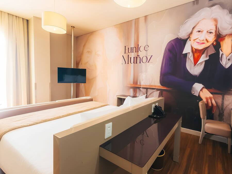 A hotel room with a bed, a desk, a chair, a TV, and a wall with a portrait and name.