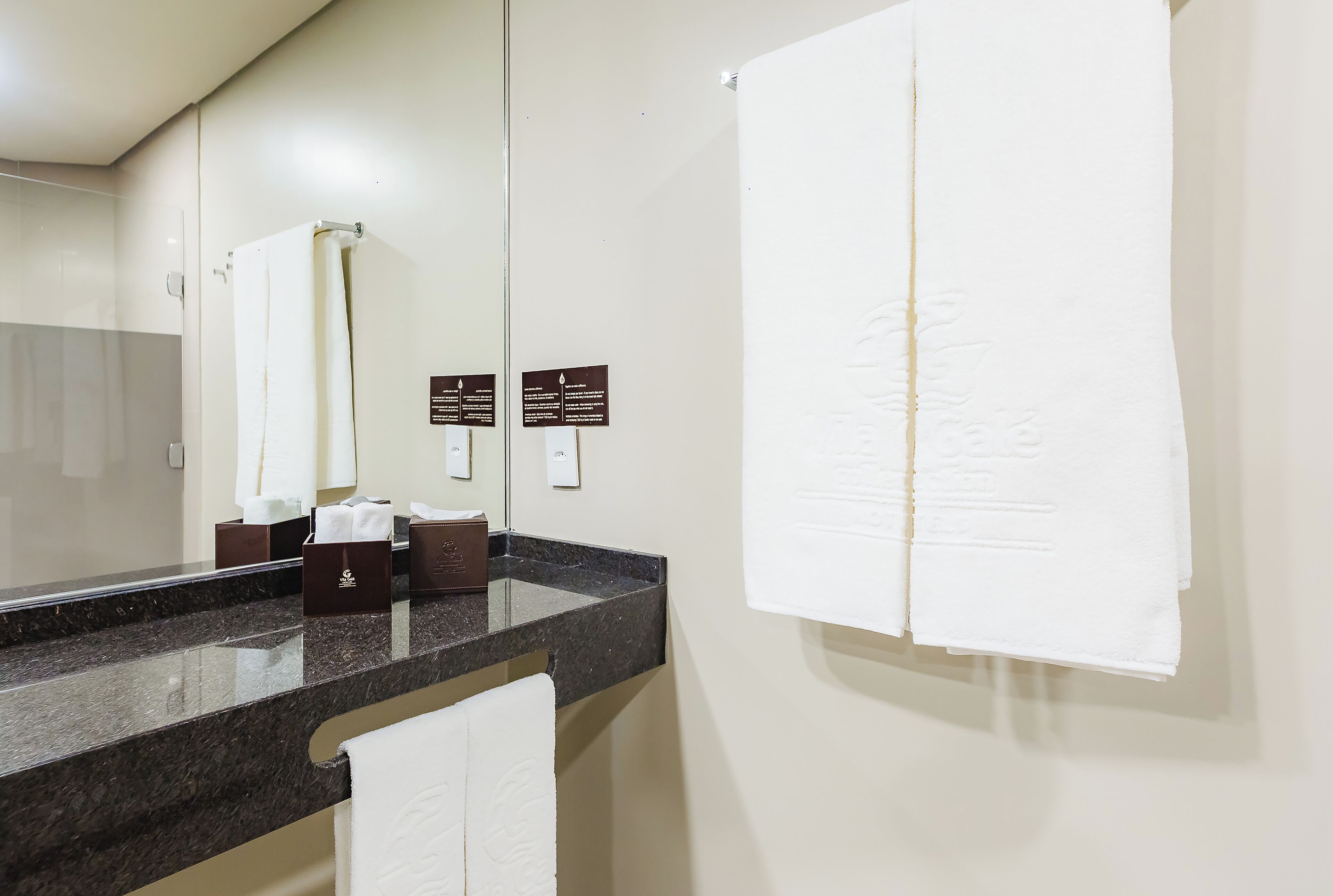 A bathroom with a marble countertop, a white towel hanging on the wall, and a mirror reflecting the towel. There are two signs on the wall, one above the mirror and one below it.