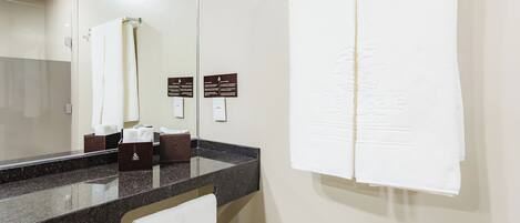 A bathroom with a marble countertop, a white towel hanging on the wall, and a mirror reflecting the towel. There are two signs on the wall, one above the mirror and one below it.