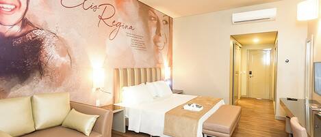 A hotel room with a large poster on the wall, a bed with white bedding, a beige sofa, a wooden floor, and a desk with a chair.