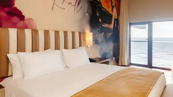 Une chambre d’hôtel avec un grand lit, une table de chevet, un mur orné d’un nom et d’un tableau, et une vue sur la mer par la fenêtre.
