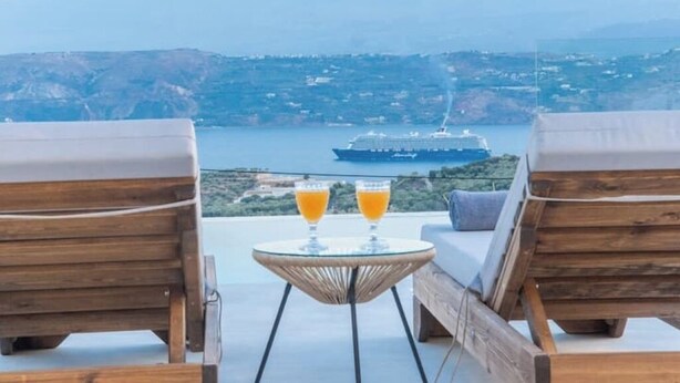 Un balcon avec deux fauteuils de jardin en bois, une petite table sur laquelle reposent deux verres de jus d’orange, et une vue sur un navire de croisière au loin.