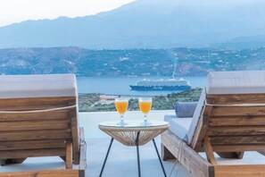 A balcony with two wooden lounge chairs, a small table with two glasses of orange juice, and a view of a cruise ship in the distance. - Fantasea Villas, villa Luna – A Peaceful Cretan Escape Above Souda Bay (Sternes)