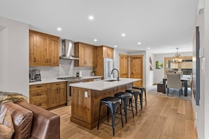 A modern kitchen with wooden cabinets, a central island with stools, and a dining area with a chandelier. - Luxury Mountain Villa | Private Hot Tub & Deck (Canmore)