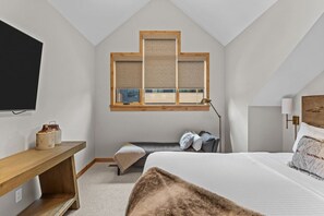 A bedroom with a sloped ceiling, a bed, a desk, a window with blinds, and a TV mounted on the wall. - Luxury Mountain Villa | Private Hot Tub & Deck (Canmore)