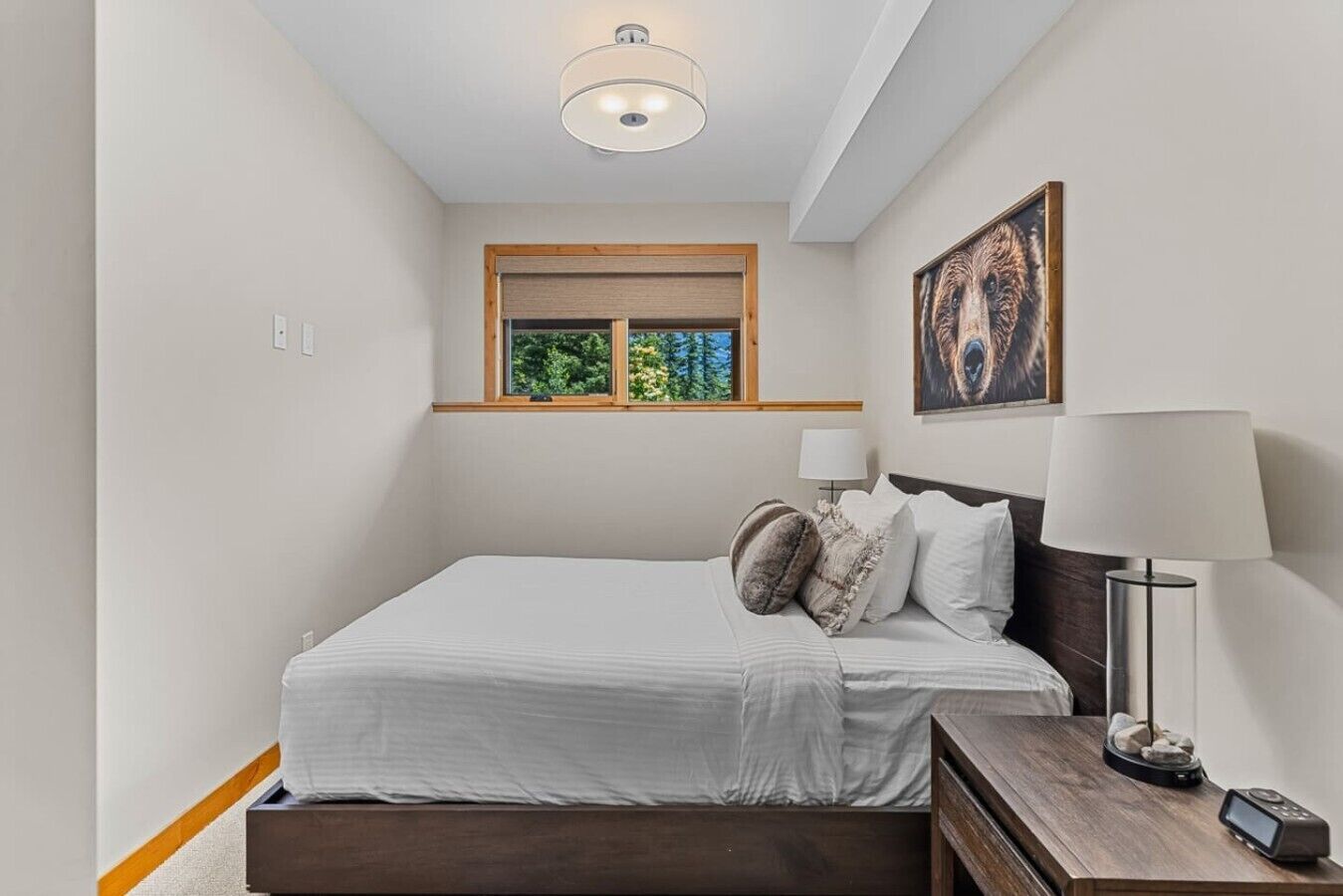 Een slaapkamer met een bed, een bedlampje, een nachtkastje, een raam met uitzicht op bomen en een schilderij aan de muur.