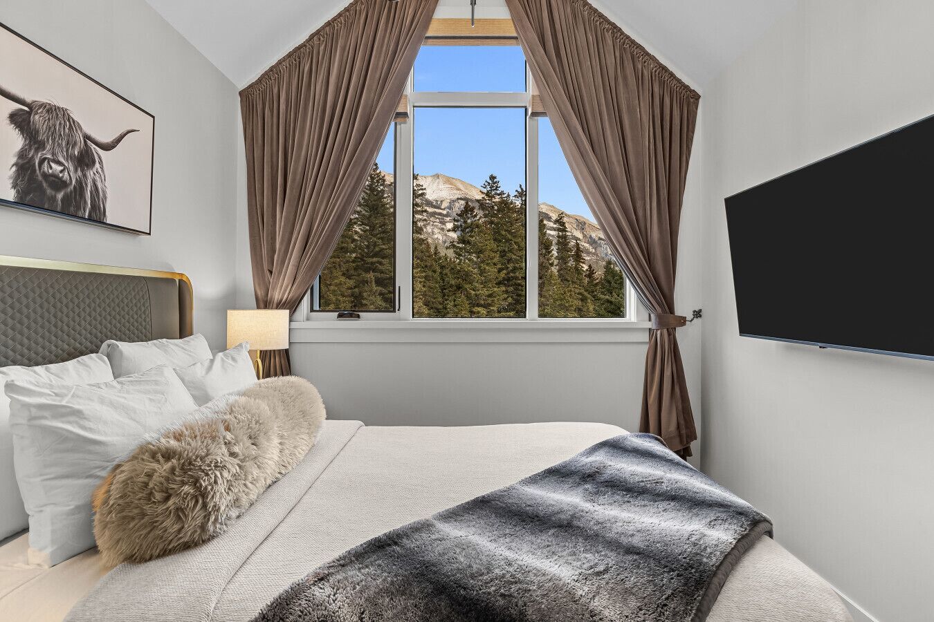 Une chambre à coucher avec un lit, une grande fenêtre avec des rideaux, un téléviseur fixé au mur et un tableau encadré accroché au mur.